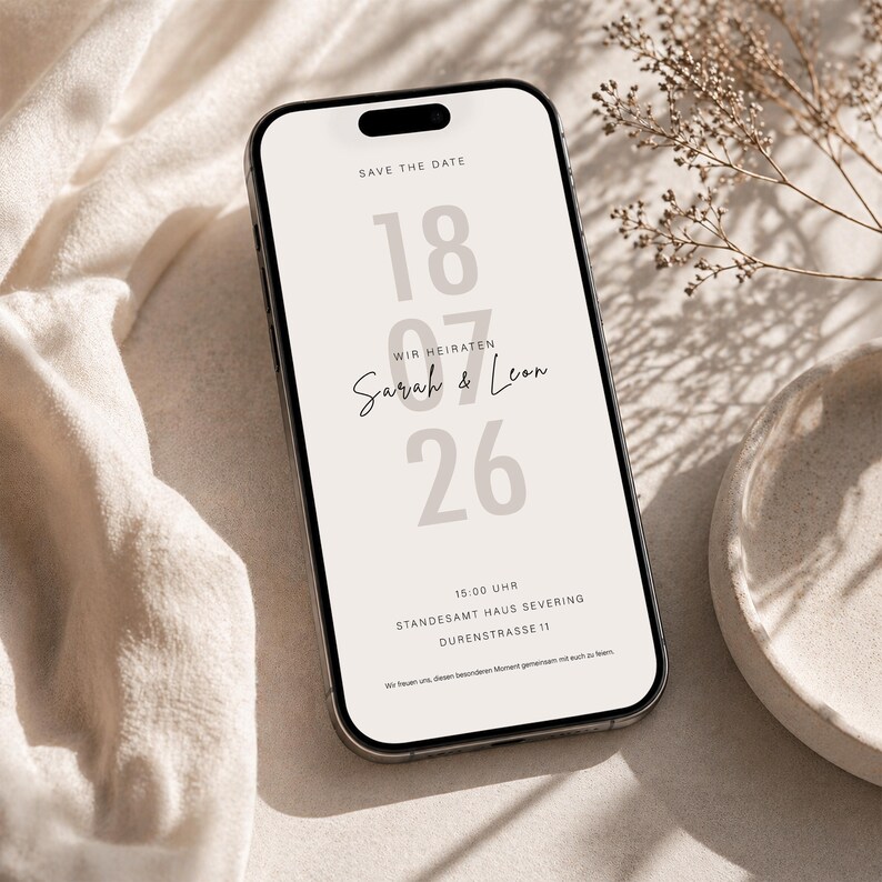May include: A smartphone displaying a digital save the date wedding invitation on a light beige background. The screen features the text SAVE THE DATE, the date 18 07 26, the names Sarah & Leon, and event details including time and location.