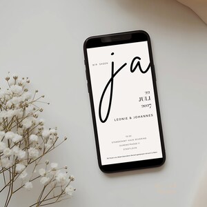 May include: A smartphone displaying a wedding invitation with the text WIR SAGEN ja, 22 JULI 2027, LEONIE & JOHANNES, 15:00, STANDESAMT HAUS SEVERING, DURENSTRASSE 11, STADTLOHN, and a small sprig of white baby's breath flowers next to the device.