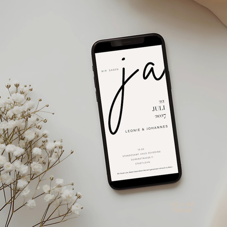 May include: A smartphone displaying a wedding invitation with the text WIR SAGEN ja, 22 JULI 2027, LEONIE & JOHANNES, 15:00, STANDESAMT HAUS SEVERING, DURENSTRASSE 11, STADTLOHN, and a small sprig of white baby's breath flowers next to the device.