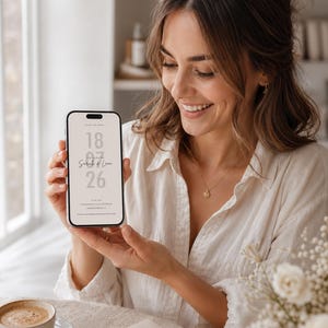 May include: A smartphone displaying a digital Save the Date announcement with the text 18 07 26 and the names Sarah & Leon in a minimalist font. The screen features a light beige background with clean, modern typography for a wedding invitation.