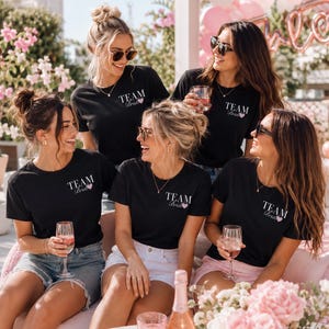 Könnte beinhalten: Eine Gruppe von Personen trägt schwarze T-Shirts mit dem Aufdruck "TEAM Bride" und einem rosa Herz. Sie lächeln und halten Gläser mit rosa Getränken. Die Szene spielt sich im Freien mit rosa Blumen und Luftballons ab.