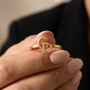 May include: A gold-toned ring featuring a polished finish and a central design element composed of the interlocking capital letters G and D. The band is smooth and circular, showcasing a metallic luster that reflects light across its surface.