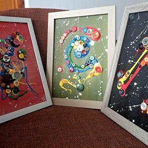 May include: Three framed abstract artworks. Each piece features colourful buttons and paint splatters on red, green, and black backgrounds. The frames are light grey, and the art is displayed on a brown surface.