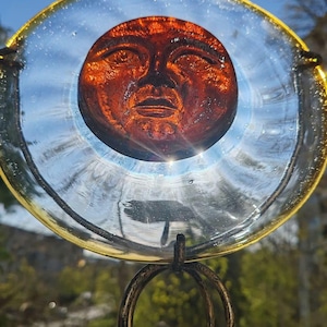 Könnte beinhalten: Ein dekorativer Glas-Sonnenfänger mit einem orangefarbenen Sonnengesicht in der Mitte. Das Sonnengesicht ist in einem klaren Glaskreis mit einem goldfarbenen Rand eingeschlossen, der von einem bronzefarbenen Metallständer getragen wird. Der Sonnenfänger ist so konzipiert, dass er Sonnenlicht einfängt.