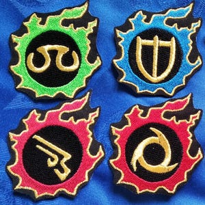 May include: Eight embroidered patches with various symbols in gold, white, and yellow on a black background. Each patch has a colored border of green, blue, or red, and a black flame-like outline. The patches are arranged on a blue fabric surface.