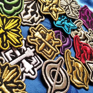 May include: A selection of embroidered patches in diverse shapes and colours, including gold, black, white, yellow, red, purple, and teal. The patches display complex designs and are arranged on a blue fabric background.
