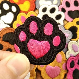 Op de afbeelding: Een verzameling kleurrijke geborduurde pootafdrukpatches. De patches hebben verschillende kleurencombinaties, waaronder zwart met roze, oranje met geel en roze met zwart. De patches zijn waarschijnlijk bedoeld voor decoratieve doeleinden, zoals op kleding of accessoires.