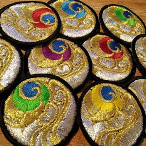Può includere: Toppe ricamate con un motivo a piuma di pavone. Ogni toppa presenta un bordo dorato, argentato e nero con un centro a spirale colorato nelle tonalità di rosso, blu, verde, giallo e viola. Le toppe sono rotonde e hanno un design dettagliato e ornato.