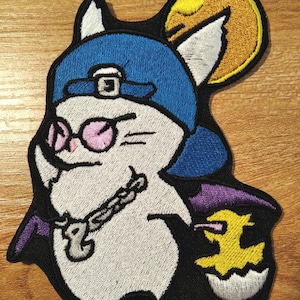 May include: Embroidered patch depicting a cartoon character wearing a blue cap and pink glasses, with a silver chain. The character has bat wings and holds a yellow chick hatching from an egg. The patch has a black border and a yellow moon.