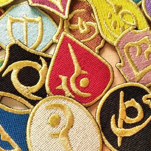 Puede incluir: Una colección de parches bordados en varias formas y colores, incluyendo rojo, azul, verde y negro. Cada parche presenta un diseño estilizado de color dorado. Los parches son probablemente para ropa o accesorios.