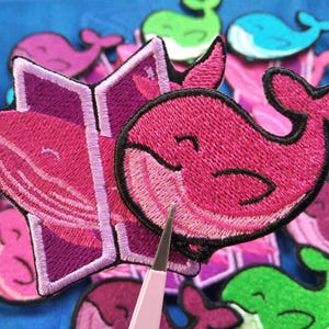 May include: Embroidered whale patches in shades of pink, purple, green, and blue. One patch is a whale reading a book. A pair of tweezers is in the foreground. The patches are on a blue background.