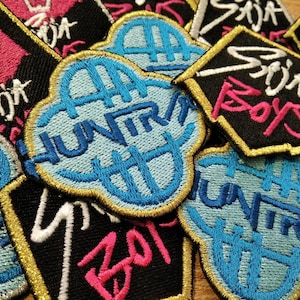 Könnte beinhalten: Eine Sammlung gestickter Aufnäher in verschiedenen Formen. Die Aufnäher zeigen Text in Weiß, Rosa und Blau mit goldenen und schwarzen Rändern. Einige Aufnäher haben eine wolkenartige Form, andere sind quadratisch oder rechteckig.