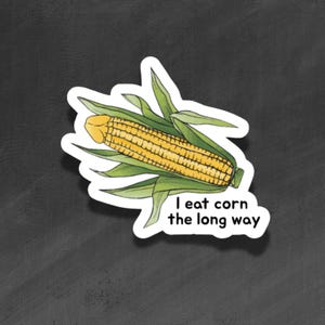 Puede incluir: Una pegatina con una ilustración de una mazorca de maíz con hojas verdes. El texto "I eat corn the long way" está debajo. La pegatina está sobre un fondo gris oscuro.