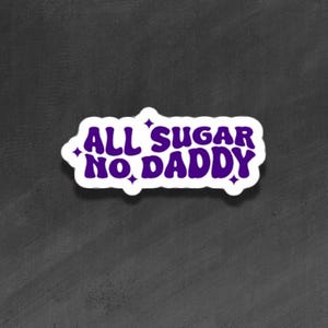 Könnte beinhalten: Ein weißer Aufkleber mit dem Text "ALL SUGAR NO DADDY" in Lila, weiß umrandet. Der Aufkleber hat eine Retro-Schriftart und Sternakzente. Der Hintergrund ist eine dunkelgraue, strukturierte Oberfläche.