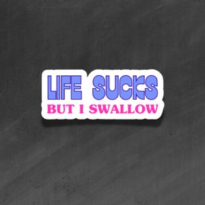Puede incluir: Una pegatina blanca con el texto "LIFE SUCKS BUT I SWALLOW" en letras azules y rosas. La pegatina tiene un fondo negro.