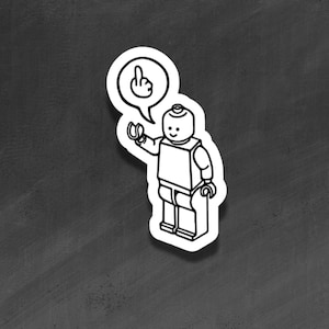 Könnte beinhalten: Ein schwarz-weißer Aufkleber mit einer Lego-ähnlichen Figur, die den Mittelfinger zeigt. Die Figur ist schwarz umrandet und weiß gefüllt, und eine Sprechblase darüber zeigt ebenfalls den Mittelfinger. Der Aufkleber befindet sich auf einem dunkelgrauen Hintergrund.