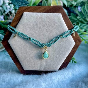 Può includere: Una collana girocollo in macramè turchese con dettagli dorati e un pendente a goccia. La collana è esposta su una superficie grigio chiaro all'interno di una cornice esagonale in legno, mettendo in risalto l'intricato lavoro di annodatura e il design.