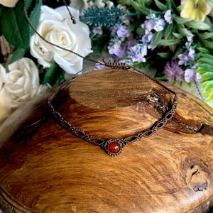 Peut inclure: Un collier ras-du-cou en macramé fait à la main, orné d'une pierre rouge centrale. Le cordon marron foncé est noué de manière complexe, créant un motif délicat. Le collier est présenté sur une surface en bois, entouré de fleurs.