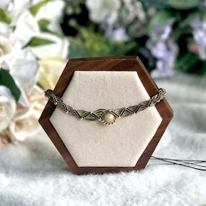 May include: A macrame necklace with a central pale green stone and gold beads. The necklace is displayed on a wooden hexagonal stand with a cream interior. The background features white flowers and greenery, creating a natural aesthetic.