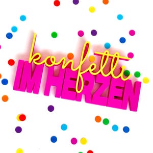 Könnte beinhalten: Ein dekoratives Schild mit dem gelben Schreibschrift-Text konfetti, der über dem kräftigen, pinkfarbenen Blockschrift-Text IM HERZEN liegt, umgeben von verstreuten, bunten, kreisförmigen Konfettipunkten in Blau, Grün, Orange, Violett und Rot.