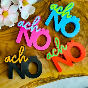 Könnte beinhalten: Eine Sammlung farbenfroher, 3D-gedruckter Schilder mit der Aufschrift "ach NO". Die Schilder sind in verschiedenen Farben gehalten, darunter Pink, Blau, Orange und Schwarz, mit kontrastierenden Akzentfarben. Die Schilder sind auf einer Holzoberfläche mit Blumen angeordnet.