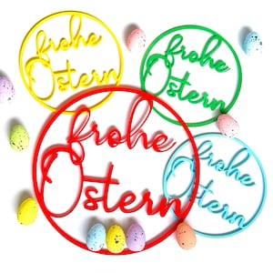 Könnte beinhalten: Vier farbenfrohe kreisförmige Dekorationen mit dem deutschen Schriftzug "Frohe Ostern" (Frohe Ostern) in einer Schreibschrift. Die Kreise sind gelb, rot, grün und blau. Gefleckte Ostereier in verschiedenen Farben umgeben die Dekorationen.