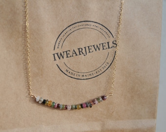 Pode incluir: Um delicado colar de ouro com uma fileira curva de contas de pedras preciosas coloridas. O colar é exibido em um saco de papel marrom com um logotipo circular que diz "I WEAR JEWELS" e "MADE IN MAINE EST 2012".