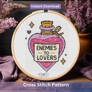 Könnte beinhalten: Ein Kreuzstichmuster in einem Holzrahmen zeigt eine rosa Zaubertrankflasche mit dem Text "ENEMIES TO LOVERS". Das Design umfasst Sterne, Herzen und die Worte "Instant Download" oben. Die Worte "Cross Stitch Pattern" stehen unten.