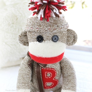May include: Handmade sock monkey toy with a brown and white knitted body, red mouth, and a red letter "B" on the chest. The monkey has button eyes and a pom-pom on its head. The text "Sock Monkey Bizz Toy Co." is visible.