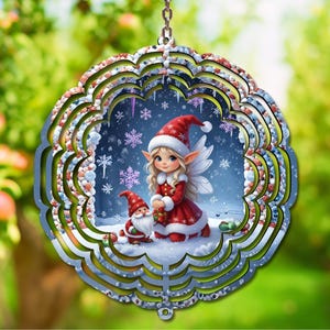 Puede incluir: Adorno navideño redondo y multicapa que presenta un hada y un gnomo en una escena nevada. El hada lleva un vestido y un sombrero rojos, mientras que el gnomo tiene un sombrero rojo y una barba blanca. El adorno tiene un marco plateado y está suspendido por una cadena.