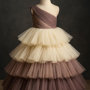 Puede incluir: Vestido de tul en capas en tonos marrón y crema, exhibido en un maniquí. El corpiño es de un solo hombro con efecto degradado. La falda presenta múltiples capas de tul con volantes, creando una silueta voluminosa.