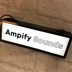 Może przedstawiać: Prostokątny podświetlany kaseton z czarną ramą i białym panelem przednim. Na froncie znajduje się napis "Ampify Sounds", gdzie "Ampify" jest zapisane pogrubioną czarną czcionką, a "Sounds" posiada czarny kontur. Z góry wychodzi czarny przewód zasilający.
