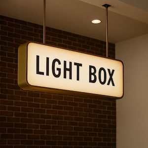 Może przedstawiać: Prostokątny szyld świetlny z napisem "LIGHT BOX" pogrubionymi czarnymi literami. Znak ma ciepłe, świecące wnętrze i złotą ramę. Wisi pod sufitem na dwóch złotych prętach.