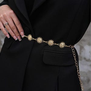Pode incluir: Um cinto de corrente dourada com pingentes em forma de sol. O cinto é usado sobre um blazer preto. Os pingentes de sol são espaçados uniformemente ao longo da corrente, e o cinto tem uma corrente pendurada. O blazer tem um design clássico.