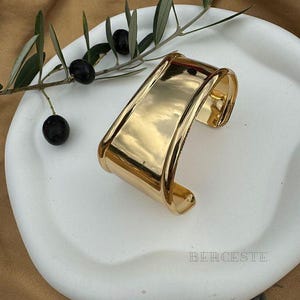 Pode incluir: Uma pulseira dourada com design retangular, repousando sobre um prato branco. A pulseira reflete a luz, e a palavra "BERCESTE" é visível. Um ramo de oliveira com azeitonas pretas adiciona um toque natural.