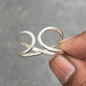 Pode incluir: Um anel prateado com um design aberto único. O anel apresenta duas formas curvas em forma de laço que estão conectadas na base. O anel é segurado pelos dedos de uma pessoa contra um fundo cinza.
