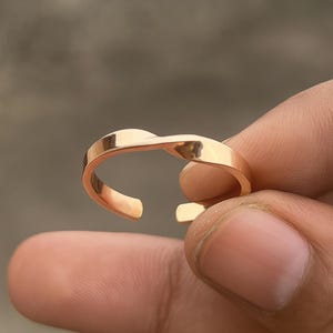 May include: A gold-colored, open-ended ring with a twisted design. The ring is made of a shiny metal and has a smooth, polished surface. The ring is held in a person's hand.