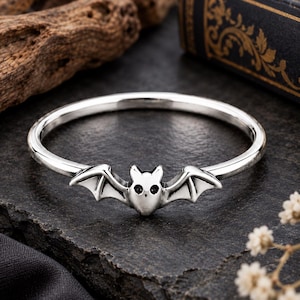 May include: A silver-toned ring featuring a central bat motif. The bat has small, dark, circular eyes and outstretched wings that integrate into the thin, polished metal band. The design is symmetrical, with a smooth, reflective finish throughout.