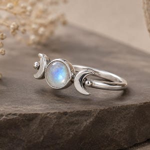 May include: A silver ring featuring a central round moonstone with a blue iridescent sheen, flanked by two crescent moon motifs on either side of the stone. The band is polished silver, and the design incorporates small spherical accents.