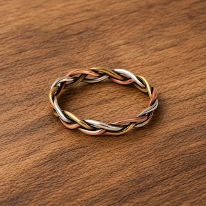 May include: A braided ring featuring interwoven strands of silver, gold, and copper-colored metal. The ring is set against a warm, brown wooden surface, highlighting the contrasting metallic hues. This piece of jewelry is a stylish accessory.