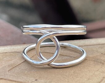 May include: A silver ring with a unique design. The ring features a double band at the top and a looped design in the center, creating an open, geometric shape. The ring is made of a shiny, reflective metal.