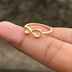 Op de afbeelding: Een goudkleurige ring met een oneindigheidssymbool. De ring heeft een eenvoudige band met het oneindigheidssymbool als middelpunt. De ring wordt op een vinger getoond.