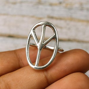 May include: A silver ring with the peace symbol design. The ring has an oval shape with the peace symbol in the centre. The ring is displayed on a person's finger.