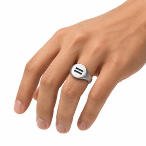 May include: A silver-toned signet ring featuring a circular face with a cut-out horizontal double-line symbol. The ring has a smooth, matte metallic finish and a solid band that tapers slightly toward the back, worn on the ring finger.