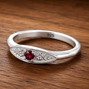 May include: A silver ring with a 925 hallmark stamped on the inner band. The ring features a central marquise-shaped setting containing a round red gemstone flanked by two small engraved floral motifs with leaf patterns on a textured background.