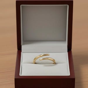 Puede incluir: Un anillo dorado con un diseño abierto único, presentado en una caja de joyería de madera oscura con un interior gris claro. Las líneas suaves y curvas del anillo crean una estética elegante y moderna.