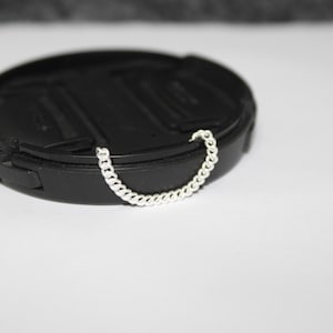 May include: A silver chain attached to a black camera lens cap. The chain is made of small, interconnected links. The lens cap is round and has a matte finish.