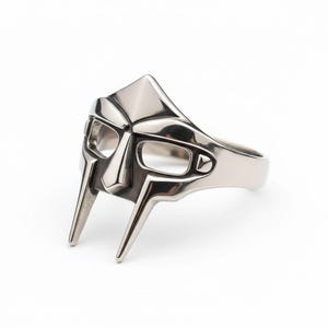 May include: A silver-toned metal ring featuring a stylised gladiator mask design. The ring has a polished finish with cut-out eye sockets and two downward-pointing prongs extending from the mask, forming the band that wraps around the finger.