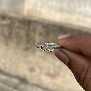 May include: A silver ring with a decorative design. The ring features a stylized, symmetrical pattern of curved lines. The ring is held in a hand against a blurred background.