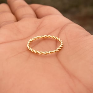 Op de afbeelding: Een goudkleurige ring met een gedraaid touwontwerp. De ring is een eenvoudige band en het gedraaide patroon geeft hem een ​​gestructureerd uiterlijk. De ring wordt op de handpalm van een persoon getoond.